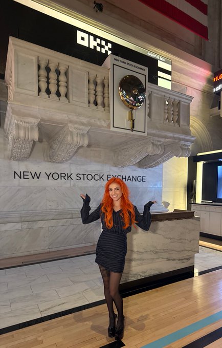 A woman with bright orange hair poses in front of the iconic New York Stock Exchange (NYSE) bell and facade, pointing toward the golden bell.
