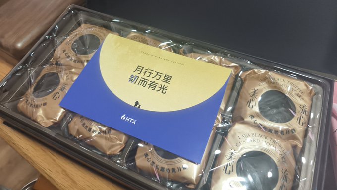 A black tray holds individually wrapped truffles with a yellow and blue card reading '月行万里 韧而有光' and 'HTX', suggesting a corporate gift from HTX.