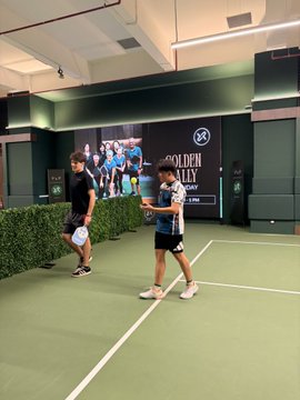 Two men stand on an indoor green tennis court, with a large 'GOLDEN PLAY' banner on the wall behind them.