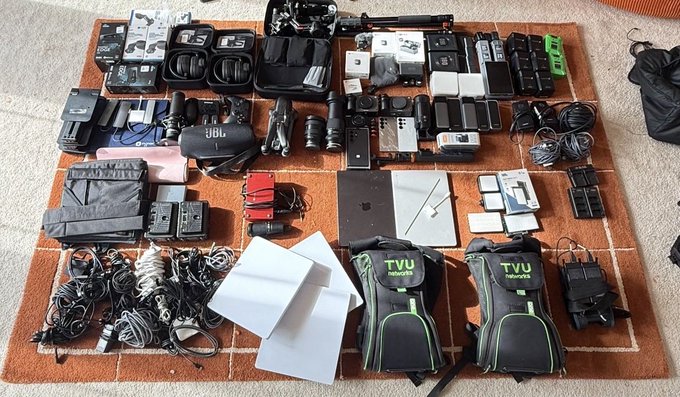 A comprehensive "One Man Prod #ARK" setup is laid out on a rug, featuring two TVU backpacks, a JBL speaker, multiple cameras, lenses, laptops, tablets, wireless microphones, cables, and production accessories. The gear showcases a complete solo broadcast kit with live streaming and audio equipment ready for mobile production.
