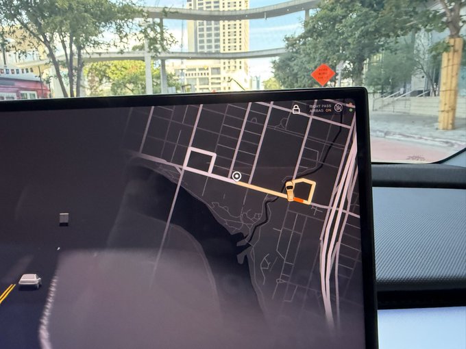 A view from inside a Tesla vehicle showing the central touchscreen display with a navigation map and a 'FRONT PASSENGER AIRBAG ON' indicator, with an urban street scene visible through the windshield.