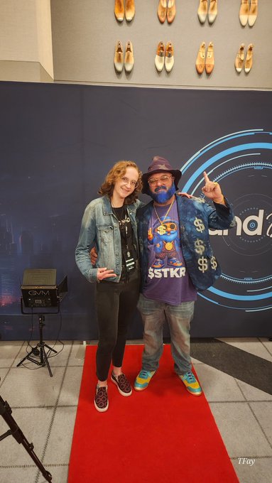 A photo of two people posing on a red carpet in front of a blue backdrop with golden shoes displayed on the wall above them.