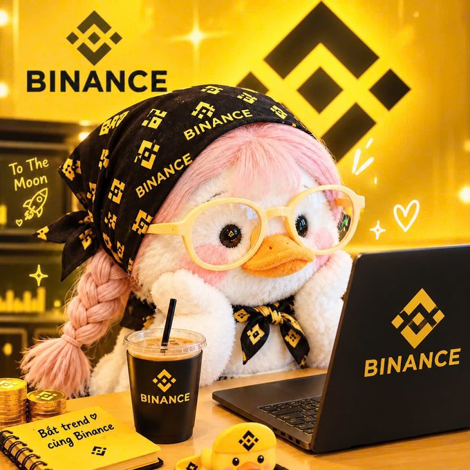 A pink plush duck toy wears a black bandana with "BINANCE" branding and a Bitcoin symbol, sitting in front of a laptop displaying the Binance logo, with a "Binance" sign in the background and a note reading "Bắt trend cùng Binance".
