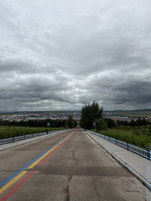 A long, empty road stretches toward a distant town under a dramatic, cloudy sky, with colorful blue, yellow, and red lines painted along its cracked pavement and white fences lining the sides. The image of a distant journey contrasts with the post about taking profits on $ASTEROID and stepping away from chart-watching, as the solitary road visually echoes the user's brief departure from the market to handle everyday tasks like waiting at a laundromat.