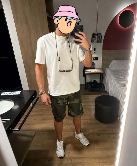 The image shows a person in a hotel room wearing a white t-shirt, camouflage shorts, and sneakers, with sunglasses hanging from their shirt. This visual aligns with the post's casual tone and implies a traveler's presence, possibly indicating the person's arrival or return.