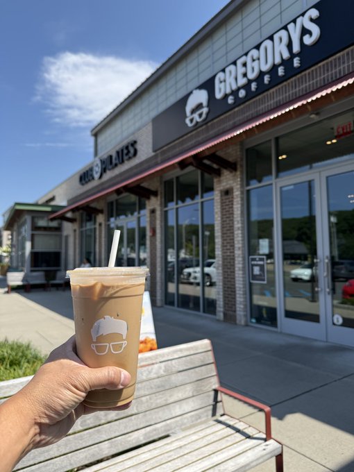 A hand holds an iced coffee drink in a clear plastic cup with the Gregorys Coffee logo, with the storefront (including 'CHOPPLATES' next door) in the background.