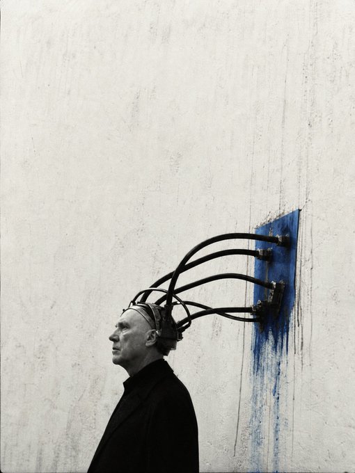 A surreal black-and-white photograph showing a man in a dark coat whose brain cables extend out and plug into a blue port on a white concrete wall, with blue paint dripping down.