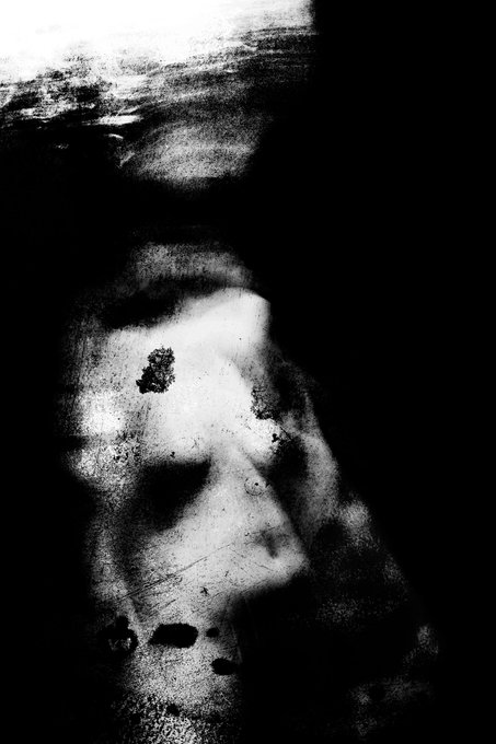A grainy, black-and-white self-portrait shows a faint, ghostly face emerging from deep shadows, characterized by high contrast and a textured, scratched appearance.