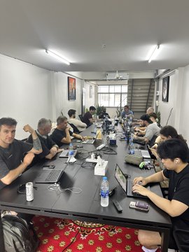 A group of people sit at a long conference table in a brightly lit room, working on laptops with water bottles and papers scattered around.
