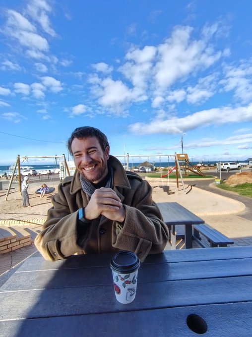 A man smiles warmly at a picnic table with a coffee cup, set against a scenic coastal backdrop with a playground and blue sky. The cheerful seaside morning perfectly matches the upbeat, optimistic tone of the "Good morning! This is gonna be a great week" message.