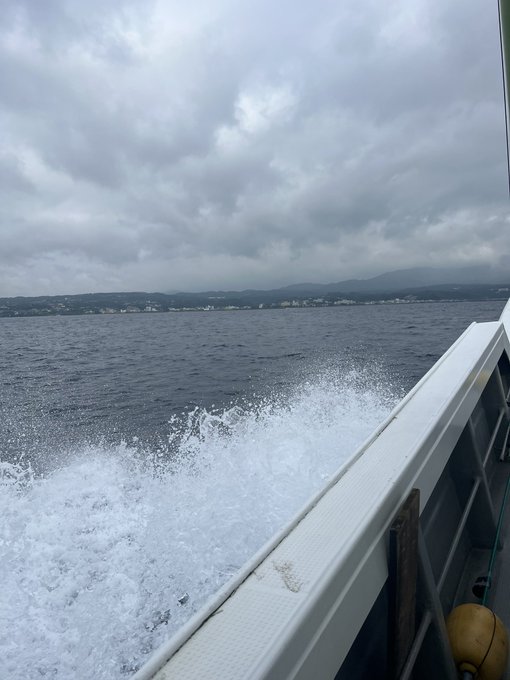 The image shows a boat cutting through choppy water, creating white waves that are crashing against its side. The sky above is cloudy, adding to the dramatic atmosphere of the scene. In the background, there's a city visible at the edge of the water. The caption on the image reads "#stepn #おみそん". This could be a reference to a specific location or event in the area where the boat is passing through.