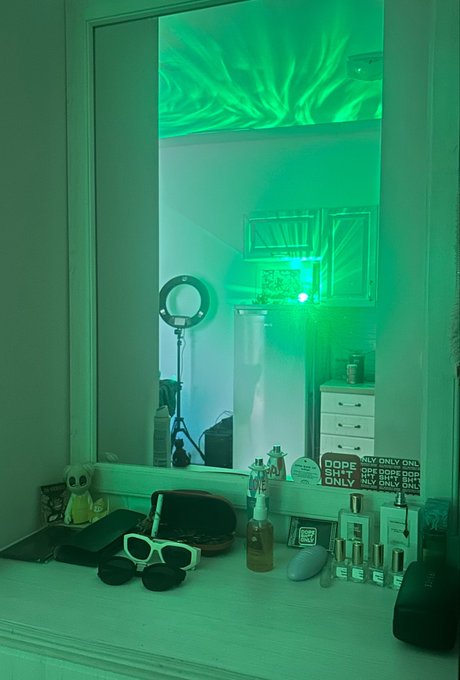A vanity mirror bathed in eerie green light displays a desk cluttered with sunglasses, skincare products, and a sign reading "DOPE BUT ONLY," with the caption humorously tying the supernatural green glow to a desperate crypto trader's attempt to summon a bull market.