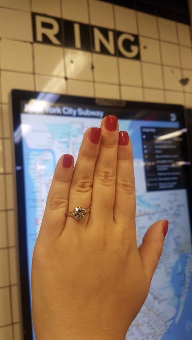 A hand displays an engagement ring with a diamond solitaire, set against the backdrop of a New York City Subway map at the Spring Street CE station. The iconic "RING" sign above the map creates a fitting coincidence for the engagement announcement.
