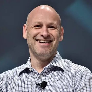 The image features a man wearing a blue and white checkered shirt with a black microphone around his neck, smiling at the camera. The background of the image is dark, which contrasts with the bright smile on the man's face. There are no other objects or texts in the image that provide additional context about the post.