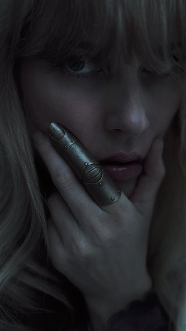 A close-up portrait of a woman with blonde hair and bangs wearing a large metallic silver ring covering her finger, set against a dark background with moody lighting.
