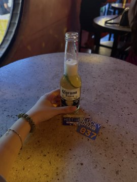 The image shows a person holding a bottle of Corona beer, which has a "corona" label on it. The person's hand is also holding a blue card with the number 037 written on it. In the background, there are two dining tables with chairs around them. One table has a bottle of beer and another one has a wallet placed on top of it.