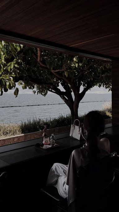A woman in a white robe sits by a window in a dimly lit room overlooking the ocean, with a tree visible outside and a tray with items on the windowsill.