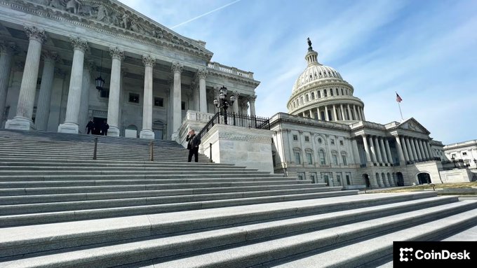 The U.S. Capitol building stands prominently under a clear blue sky, with a lone figure visible on the expansive stone steps leading to the Senate chambers. The CoinDesk watermark appears in the bottom right corner. The image visually represents the setting for the legislative efforts mentioned in the post, where Senate staff are working to reconcile the CLARITY Act between the Agriculture and Banking Committees before the August recess.