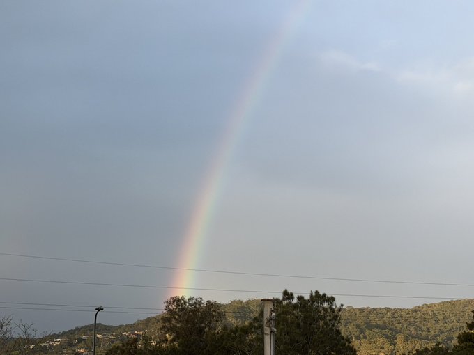 A vibrant rainbow arcs across a cloudy gray sky, descending toward a landscape of rolling hills, trees, and power lines.