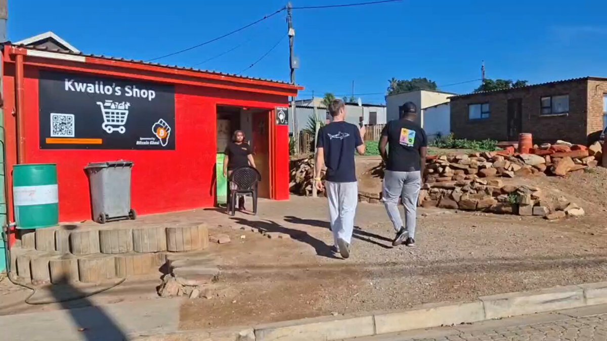 The image shows two men walking past a red building with a "Kwela's Shop" sign on it. The scene takes place outside of the shop, where there are several other people present as well. One person can be seen sitting at a table in front of the shop, while another is standing nearby. A chair and a handbag are also visible within the scene.