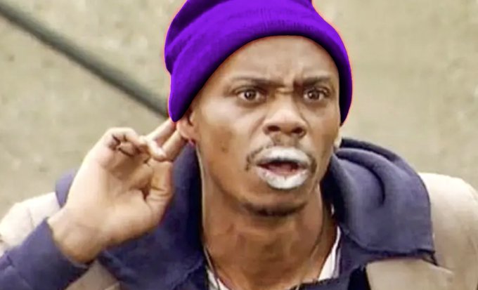 A man in a purple beanie and grey hoodie holds a finger to his ear with an exasperated expression, his mouth open in apparent frustration.