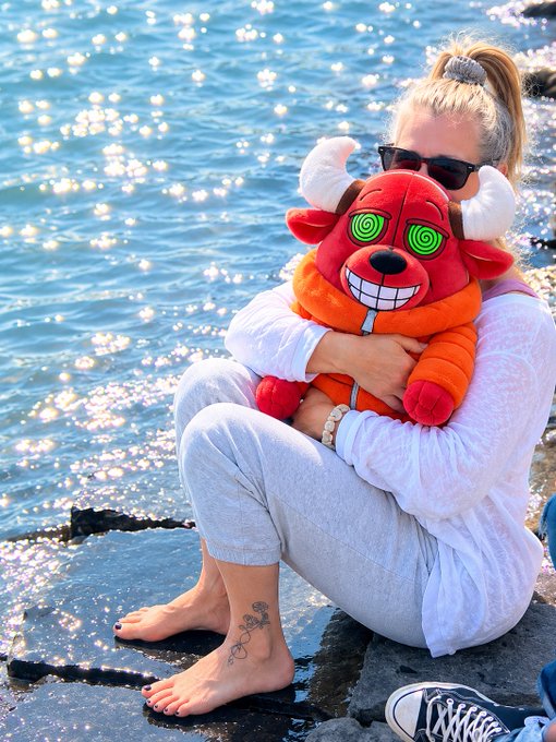A woman wearing sunglasses and a light outfit sits barefoot by sunlit water, hugging a red plush toy with horns, wide grinning teeth, and swirled green hypnotic eyes.