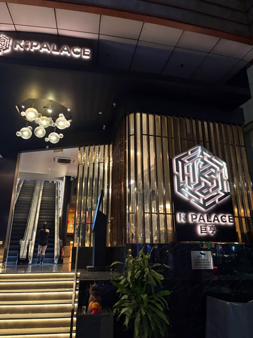 The exterior entrance of K Palace, featuring illuminated signage, an escalator, a modern chandelier, and decorative plants.