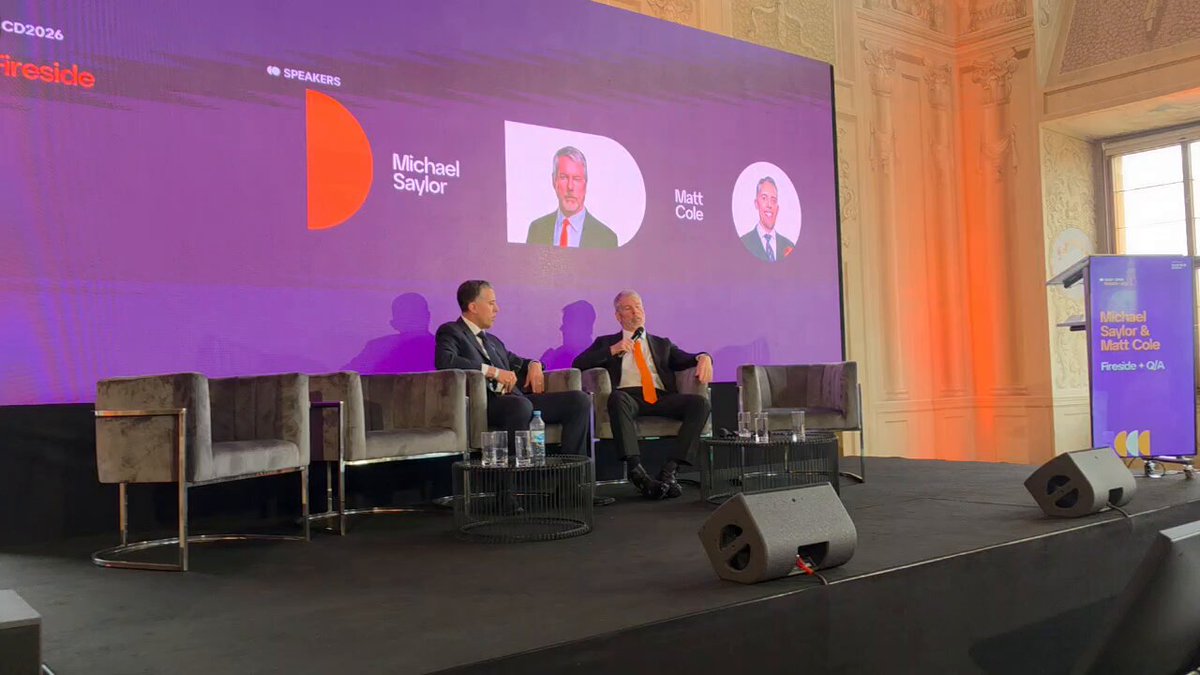 The image shows two men sitting at a table, possibly participating in a panel discussion or conference event. They are seated on chairs placed around a stage with a purple backdrop featuring three different faces of people. The man on the left side of the image is wearing a black suit and tie, while the other man has a more casual attire consisting of a gray shirt and an orange tie. The background includes a podium and a banner displaying the words "Digital Credit or Digital Money?". This suggests that they might be discussing the concept of digital currency or credit in relation to the current financial landscape.