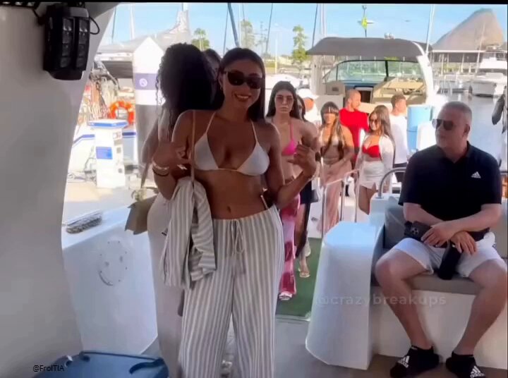 The image shows a group of people standing together, possibly at a party or gathering. One woman stands out as she wears a bikini top and striped pants while holding her purse. The other women are dressed more casually, with some wearing swimsuits. A man is sitting on a bench nearby, observing the scene. In the background, there are boats visible in the water, suggesting that this event might be taking place near a marina or waterfront location.