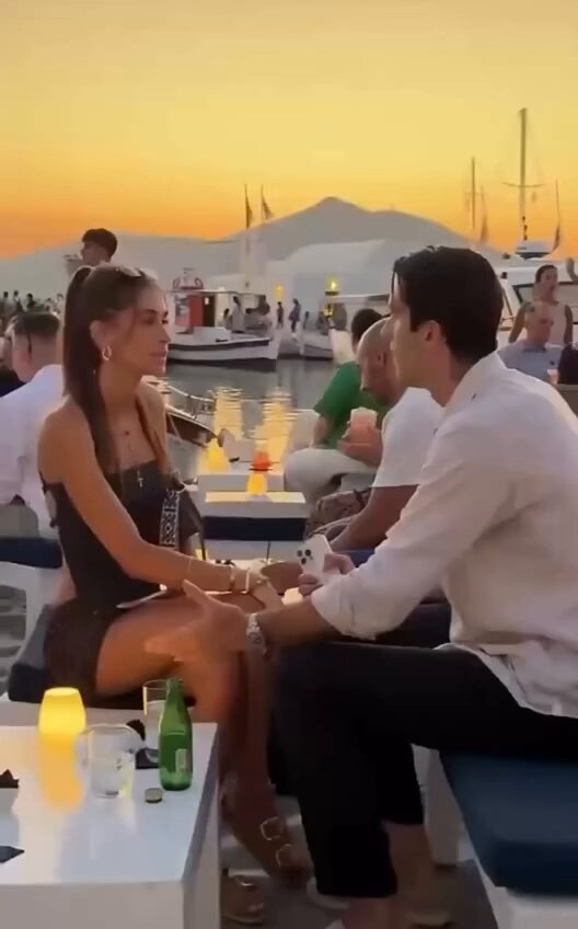 The image shows a man and woman sitting at a table with drinks, including beer bottles and wine glasses. They are engaged in conversation while enjoying their time together. The scene takes place near a body of water where boats can be seen in the background. The photo appears to have been taken during sunset or dusk, as suggested by the warm colors in the sky.