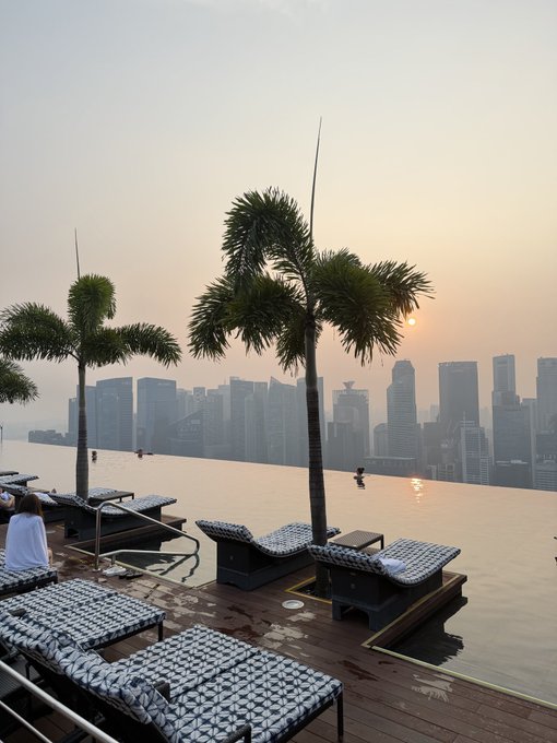 An infinity pool overlooking a hazy sunset skyline with palm trees and patterned lounge chairs, capturing a moment of in-person connection after a crypto event. The serene view reflects the post's sentiment of taking a brief reprieve to recharge before diving back into focused work.