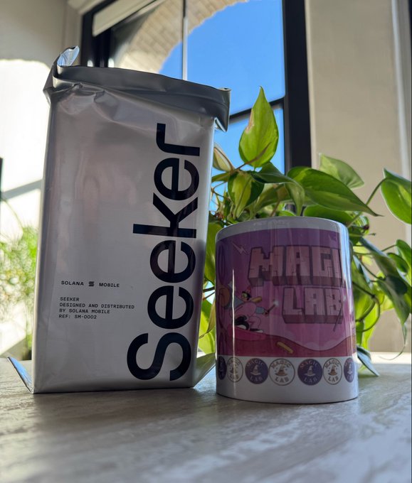A silver 'Seeker' package by Solana Mobile sits next to a cylindrical 'Magi Lab' container with a leafy plant behind them on a windowsill.