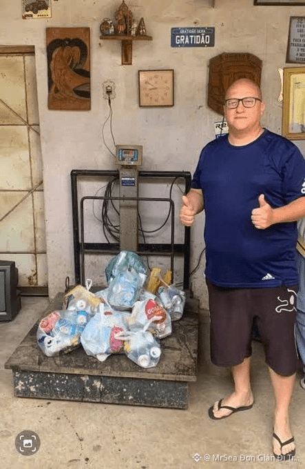 A man in a blue t-shirt and black shorts giving two thumbs up while standing next to a scale loaded with multiple bags of recyclable plastic bottles, with a 'MR SEA BEN GIAN' watermark.