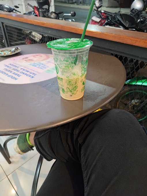 An iced beverage in a plastic cup with green graphics and a green straw sits on a round café table, with motorcycles visible parked on the street in the background.