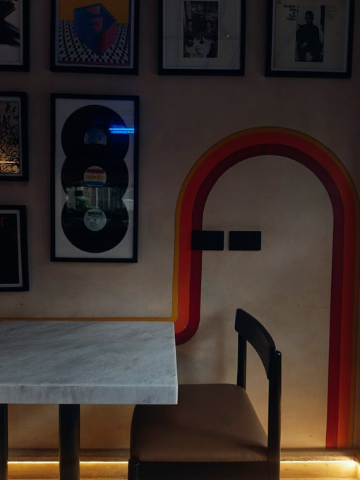 A dimly lit interior scene showing a marble-topped table and a brown leather chair positioned in front of a wall decorated with framed vinyl records and pictures, alongside a distinctive arched doorway with red and yellow trim.