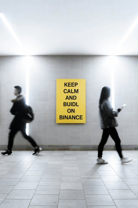 Two blurred figures walk past a yellow poster on a tiled wall that reads 'KEEP CALM AND BUIDL ON BINANCE' in a subway or underground passage.