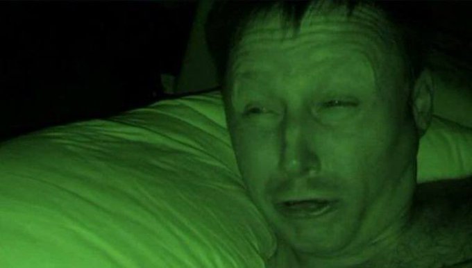A green-tinted, low-light close-up shows a middle-aged man resting his head on a white pillow with a tired or distressed expression.