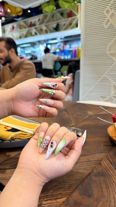 Two hands displaying elaborately decorated stiletto nails with pastel colors, patterns, and 3D embellishments, set in what appears to be a restaurant or bar setting.