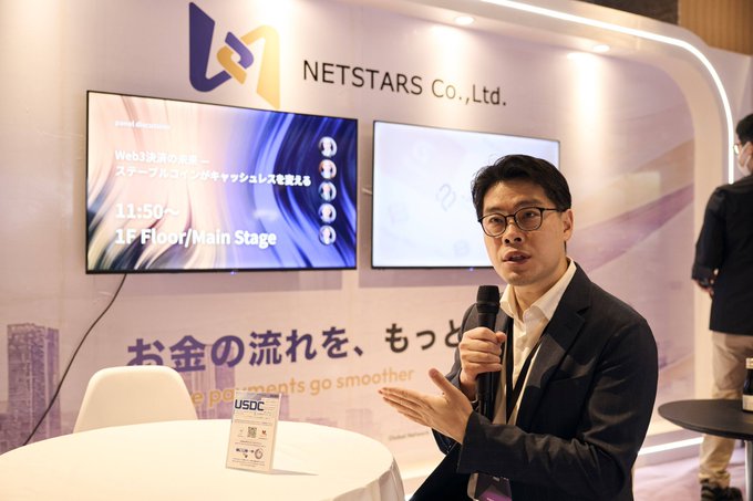 A man in glasses and a dark suit speaks into a microphone at the NETSTARS Co., Ltd. booth at USDC, with screens displaying "Web3決済の未来" and Japanese text about Netstars & USDC cashless promotion.