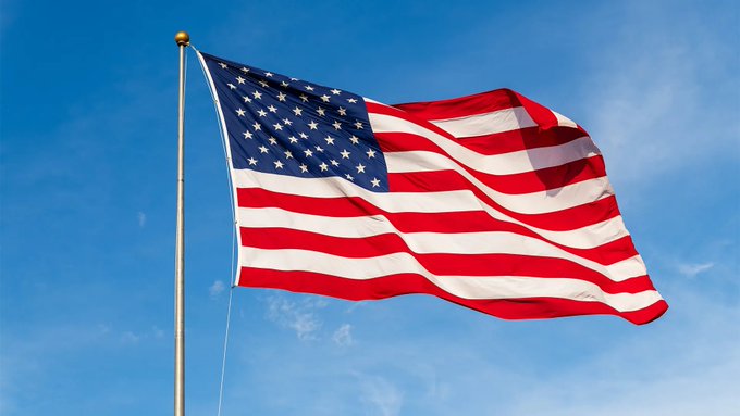 A photograph of the American flag waving against a clear blue sky on a flagpole.