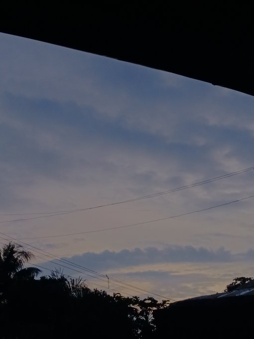 A serene photograph of a twilight sky with soft clouds, power lines stretching across the frame, and silhouetted palm trees and rooftops below.