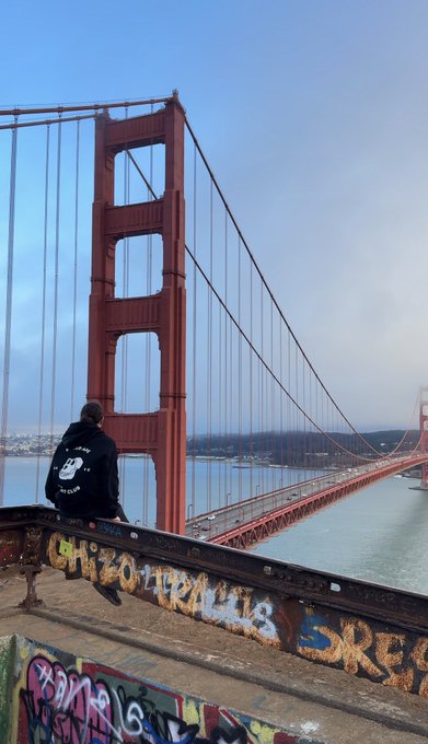 A person wearing a black hoodie sits on a graffiti-covered railing, looking out at the Golden Gate Bridge shrouded in mist under a partly cloudy sky.