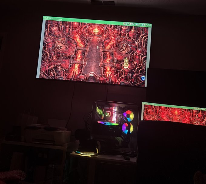 A dimly lit gaming setup with a wall-mounted monitor and a PC tower with RGB lighting, both displaying a red-themed game or wallpaper.