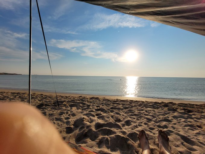 A serene beach scene viewed from under a canopy, showing sandy shores, calm ocean waters reflecting the bright sun, and a person's feet and legs visible in the foreground.