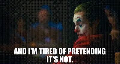 The Joker with green hair and clown makeup, wearing a red coat, smiles against a blurred cityscape background with white text reading 'AND I'M TIRED OF PRETENDING IT'S NOT.'