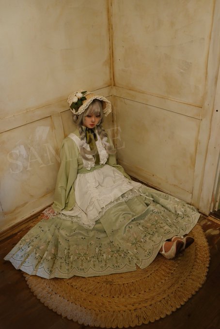 A doll with silver hair and green ribbons sits in a corner wearing an elaborate green and white dress with a matching bonnet, with the word 'SAMPLE' watermarked across the image.