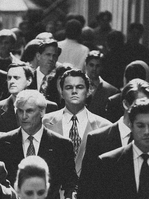 A black and white photo of Leonardo DiCaprio in a light suit walking through a crowd of men in dark suits, appearing to be a scene from a film.