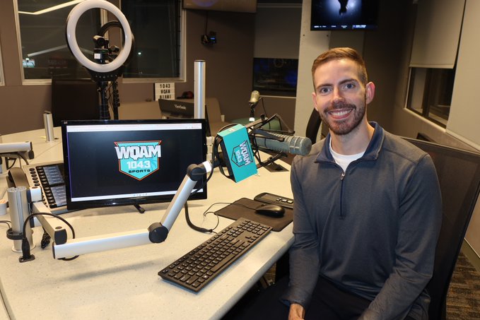 A smiling man with a beard sits at a desk in a professional radio studio, with a monitor displaying the "WOAM 104.3 Sports" logo and a ring light and microphone set up for broadcasting. The celebratory post "Congrats, Solana!" suggests this is likely the station's on-air talent marking a personal or professional milestone.