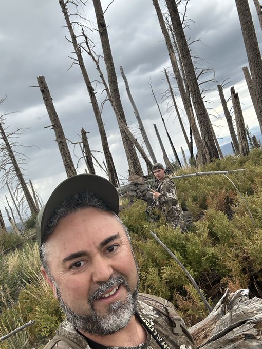 A selfie of a man with a beard wearing a cap and camouflage gear in a forested area with many dead standing trees, with another person in camouflage visible in the background.