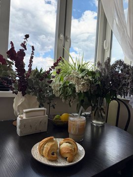 The image features a dining table with two plates of croissants placed on top. The table also has various items, including vases filled with flowers, a cup, and a bottle. In front of the table, there are two chairs, one of which is occupied by an empty chair. A window can be seen in the background, allowing natural light to enter the room. The image includes text that reads "GM my CT. Yesterday was a bit lacking in football events. All that’s left now is to wait for the Champions League final, and then the big football celebration begins".